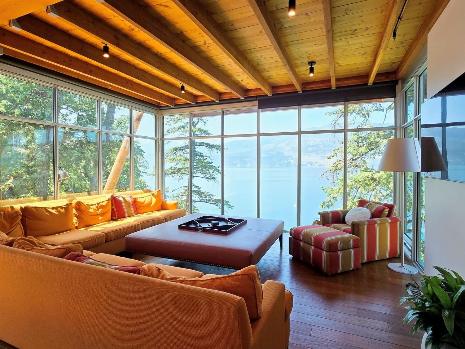 Living room with large windows, wooden ceiling, and lake view, with couches, table, and plant.