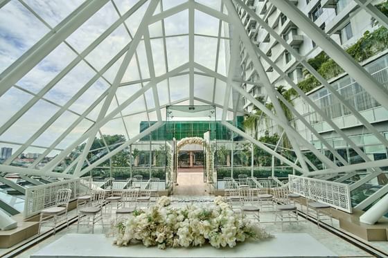 Interior of a glass pyramid wedding chapel with white chairs, floral arrangements, and city views at Vasa Hotel Surabaya
