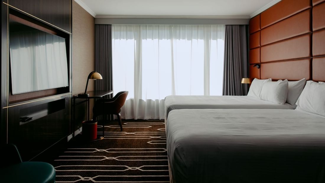 Hotel room with two beds, a desk with a lamp, and a large window with curtains.