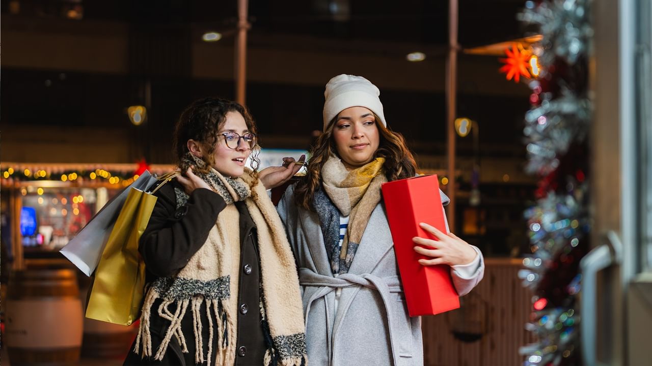 Two women with shopping bags and a red box stand in front of a decorated store.