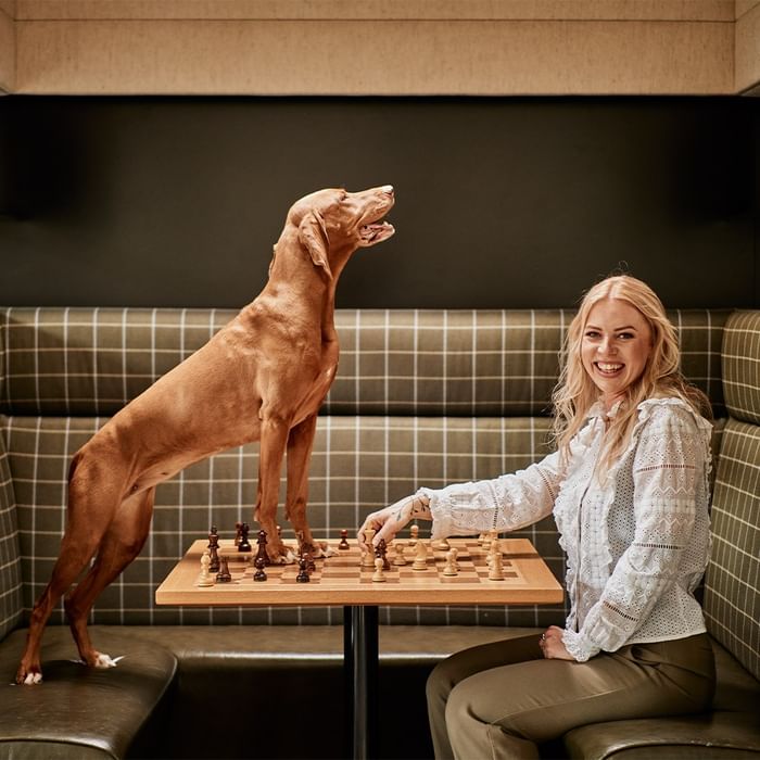 Woman and dog playing chess, dog standing on table with two front paws on board, woman smiling.