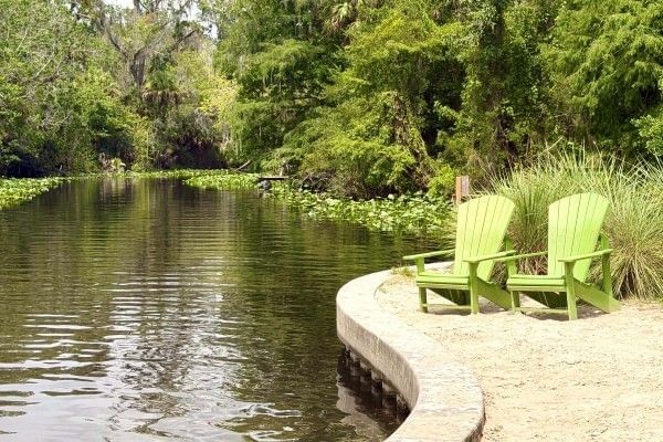 A winding river surrounded by lush greenery and two green Adirondack chairs sitting on a sandy bank at Wekiva Island in Orlando.