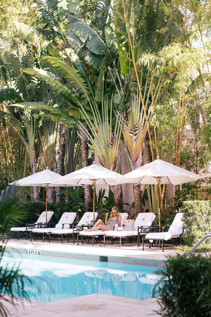 Woman in swimsuit reclining on poolside lounge chair under umbrella near palm trees at The Brazilian Court