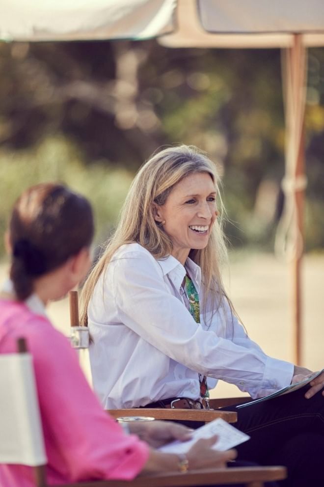 Smiling woman in a white shirt seated in a chair, chatting with a guest on a sunny beach at Marbella Club