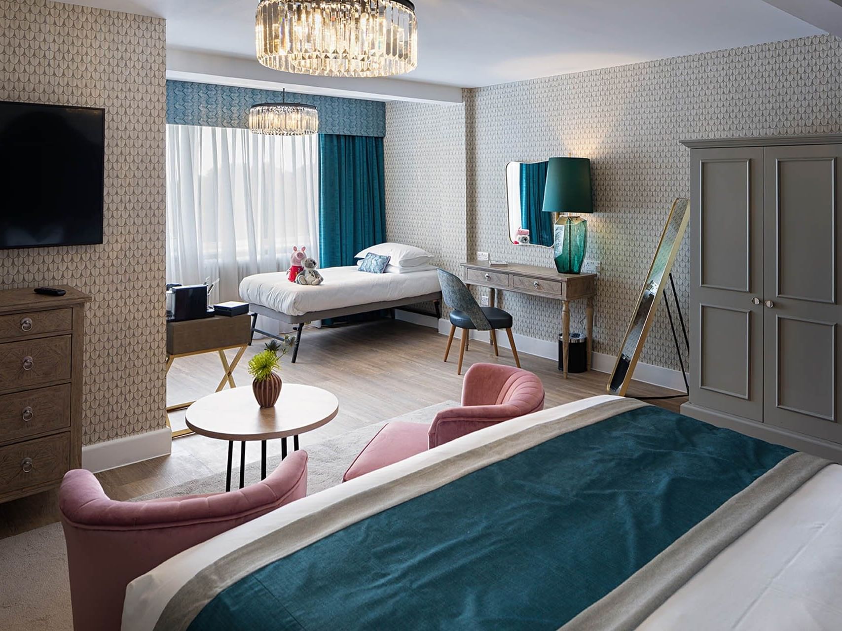 Hotel room with two beds, sitting area, chandelier, desk, mirror, and television on the wall.