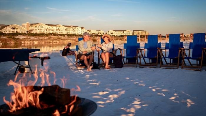 Couple by firepit on beach with blue lounge chairs lined up at sunset for Labor Day Weekend.