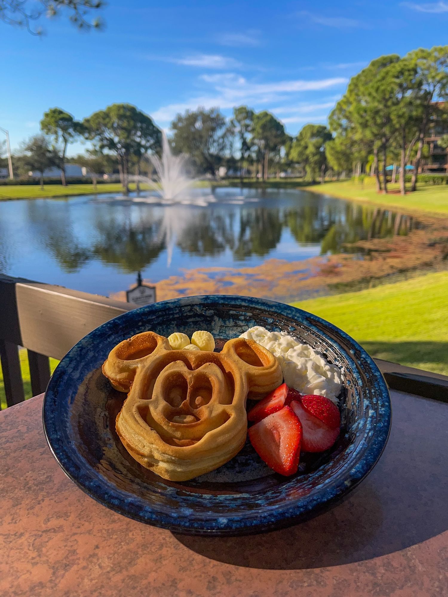 An image of a Mickey Mouse pancake on a breakfast plate on an outdoor table, with a background view of a lake with a fountain in the middle. After navigating Disney theme parks as an introvert, retreat to the serene oasis of Rosen Inn Lake Buena Vista. 