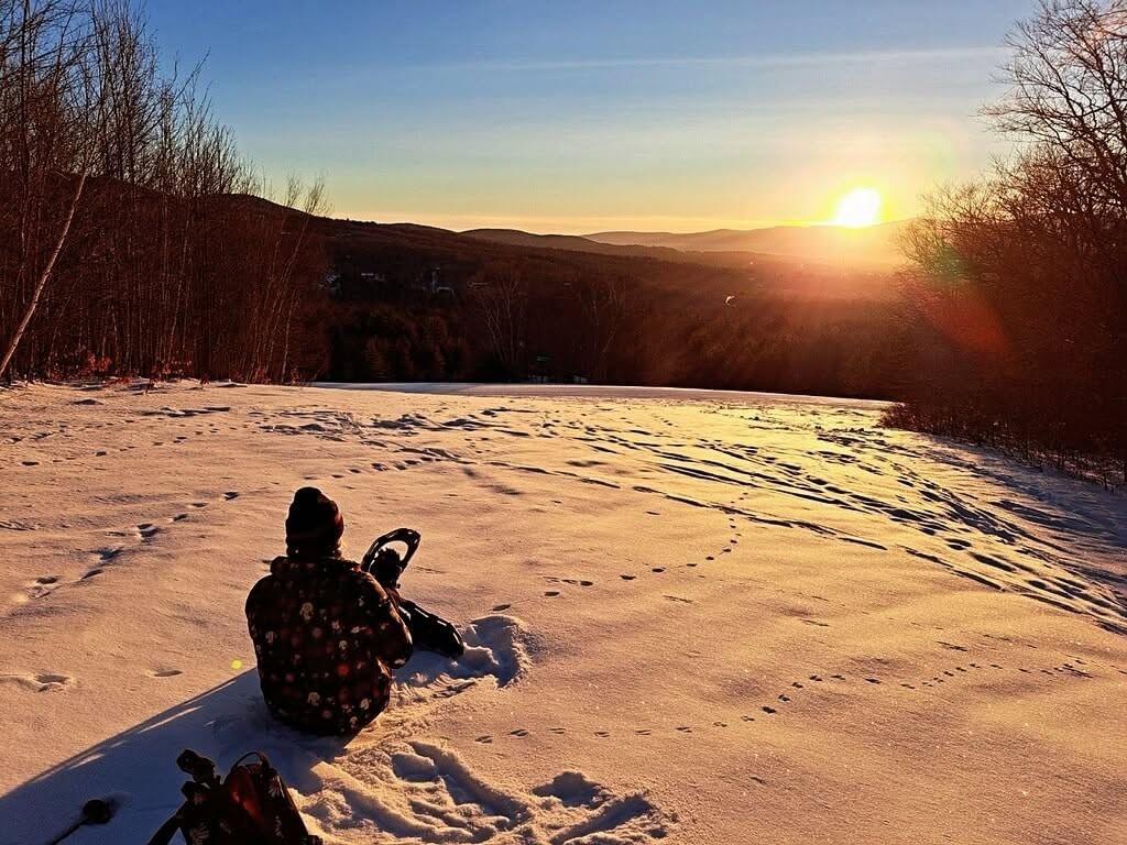Person sitting on snowy hillside watching golden sunrise over rolling mountains with Sunrise Mountain Guides.