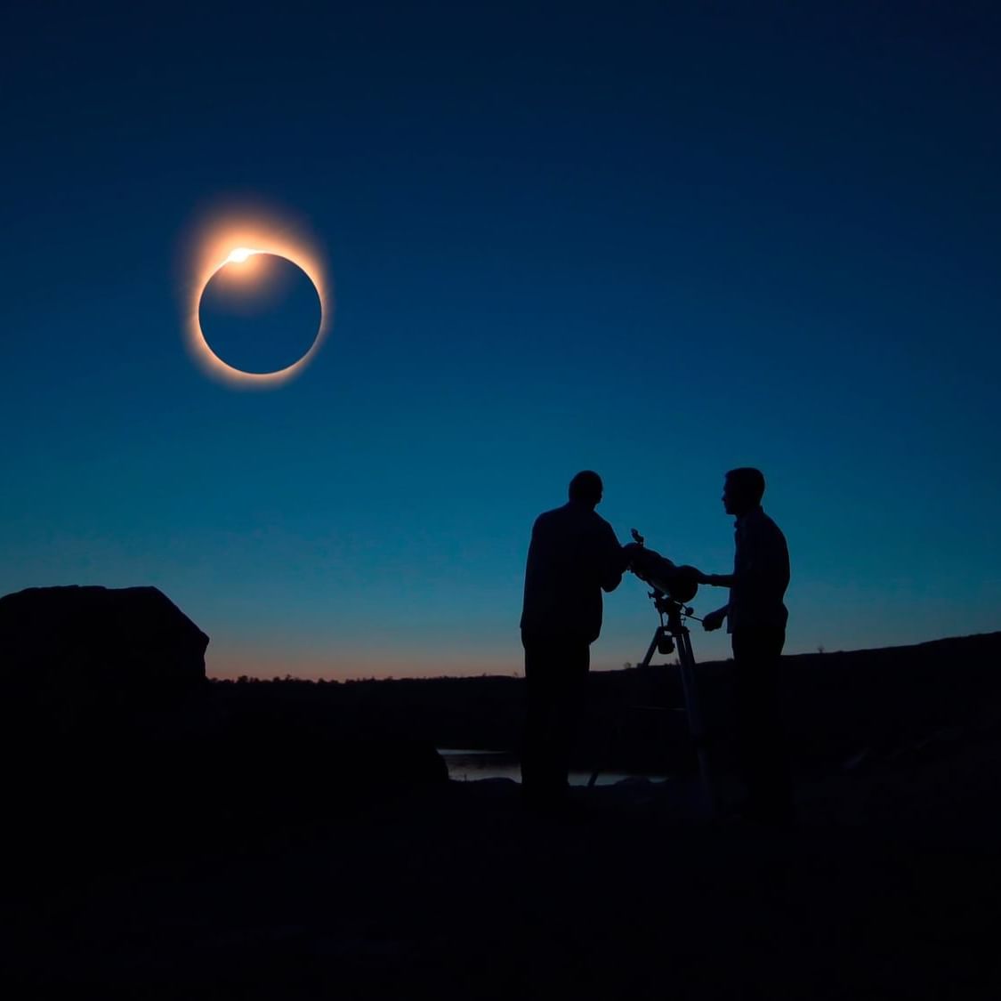 Two people with a telescope watching a solar eclipse in the evening sky.