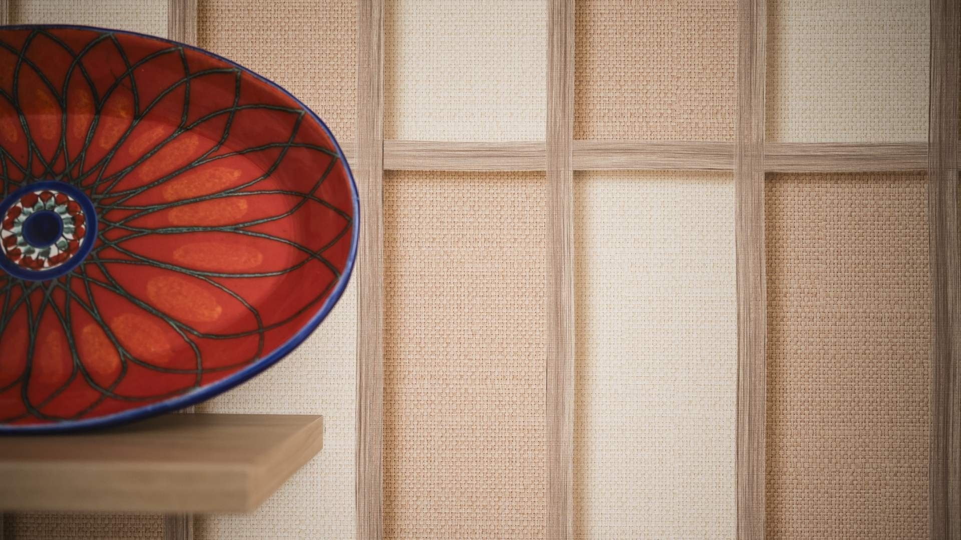 Piatto decorativo rosso e blu su una mensola di legno davanti a una parete in tessuto.