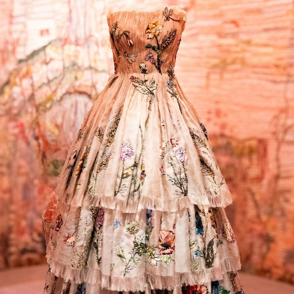 Eine ausgestellte, blumenbestickte, weiße, bodenlange Robe vor einem orangefarbenen Hintergrund.