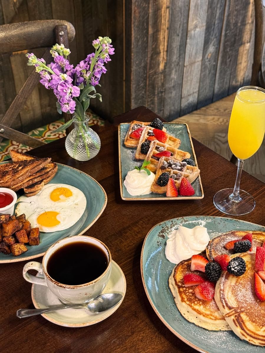 Fried eggs and waffles by a mimosa and purple flowers on a wooden table in BOTA at Warwick San Francisco