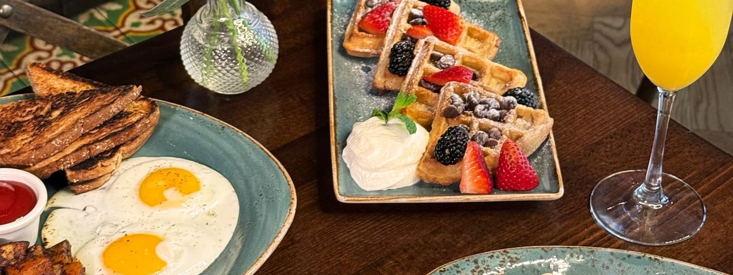 Fried eggs and waffles by a mimosa and purple flowers on a wooden table in BOTA at Warwick San Francisco