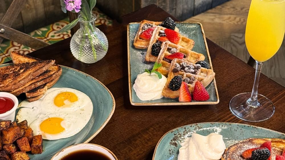 Fried eggs and waffles by a mimosa and purple flowers on a wooden table in BOTA at Warwick San Francisco