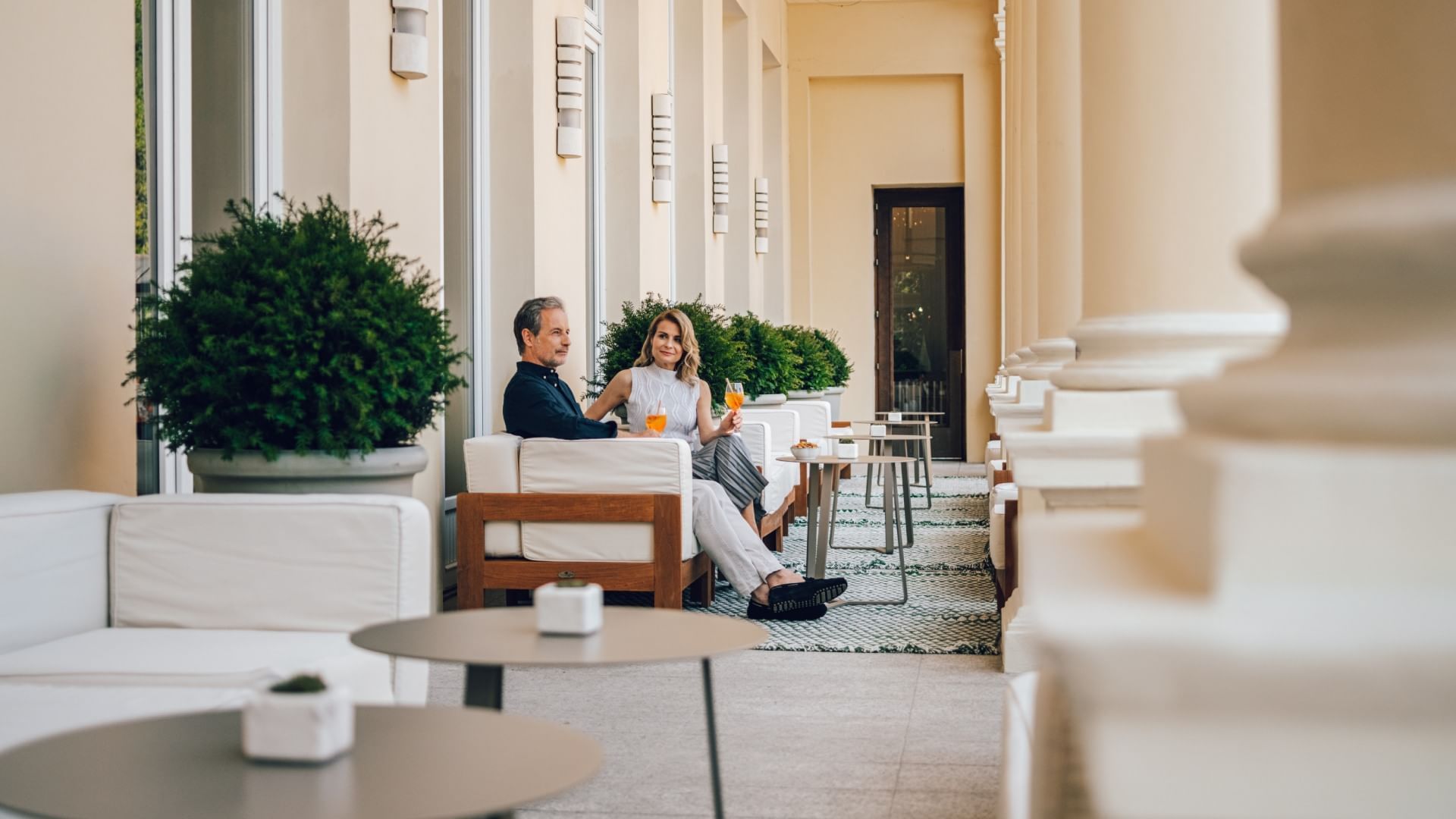 Ein Paar sitzt auf einer Terrasse, umgeben von Möbeln und Pflanzen, und genießt Getränke.