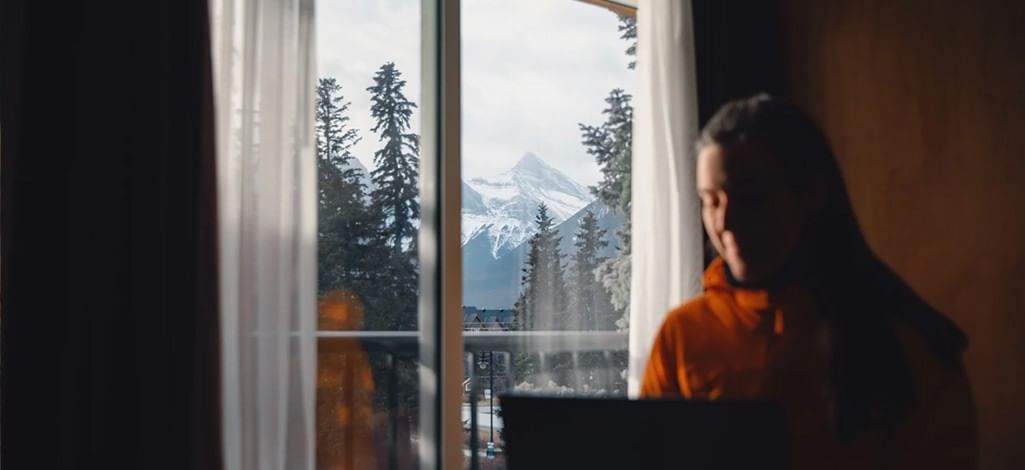 A person uses their laptop in a hotel room in Canmore with a snowy mountain view out the window.