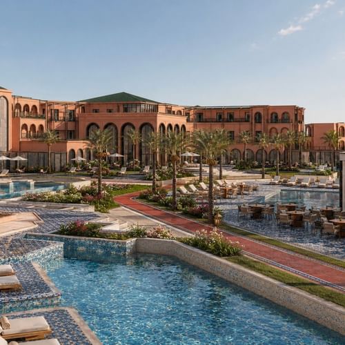 Outdoor pools with loungers and dining tables surrounded by palm trees at Palmeraie Golf Palace by Pickalbatros