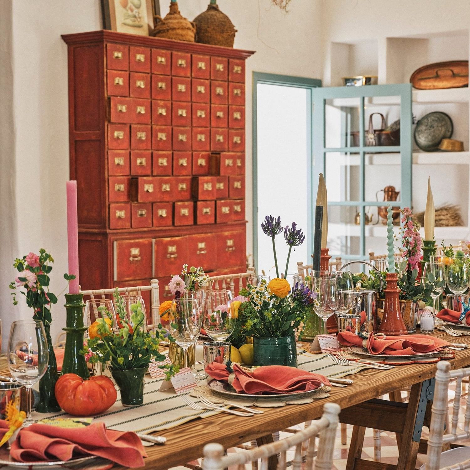 A long dining table with floral arrangements, wine glasses, and place settings in a room with a red cabinet.
