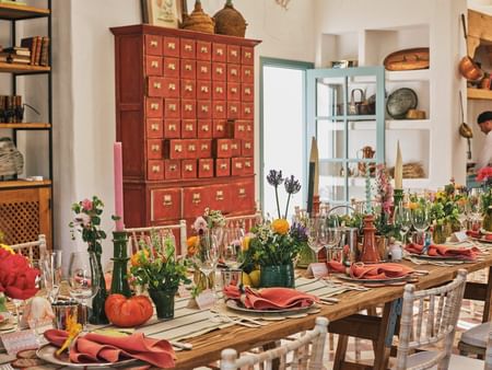 Mesa larga decorada con flores y cubiertos, junto a un armario rojo y una puerta azul.