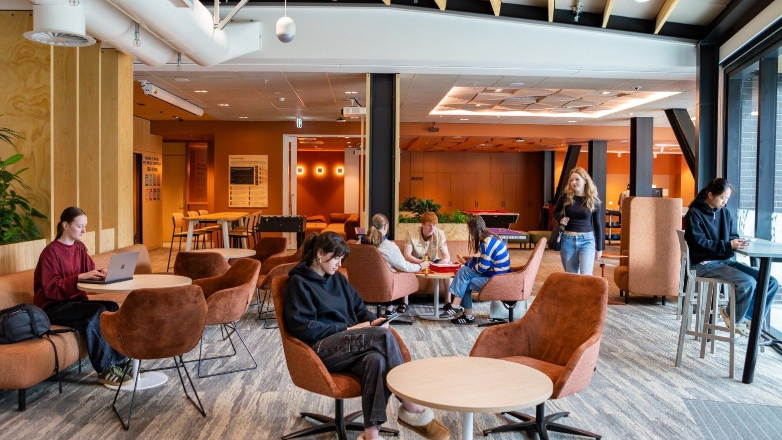 Modern communal lounge with warm wood accents, brown chairs, and guests working at Tupuārangi