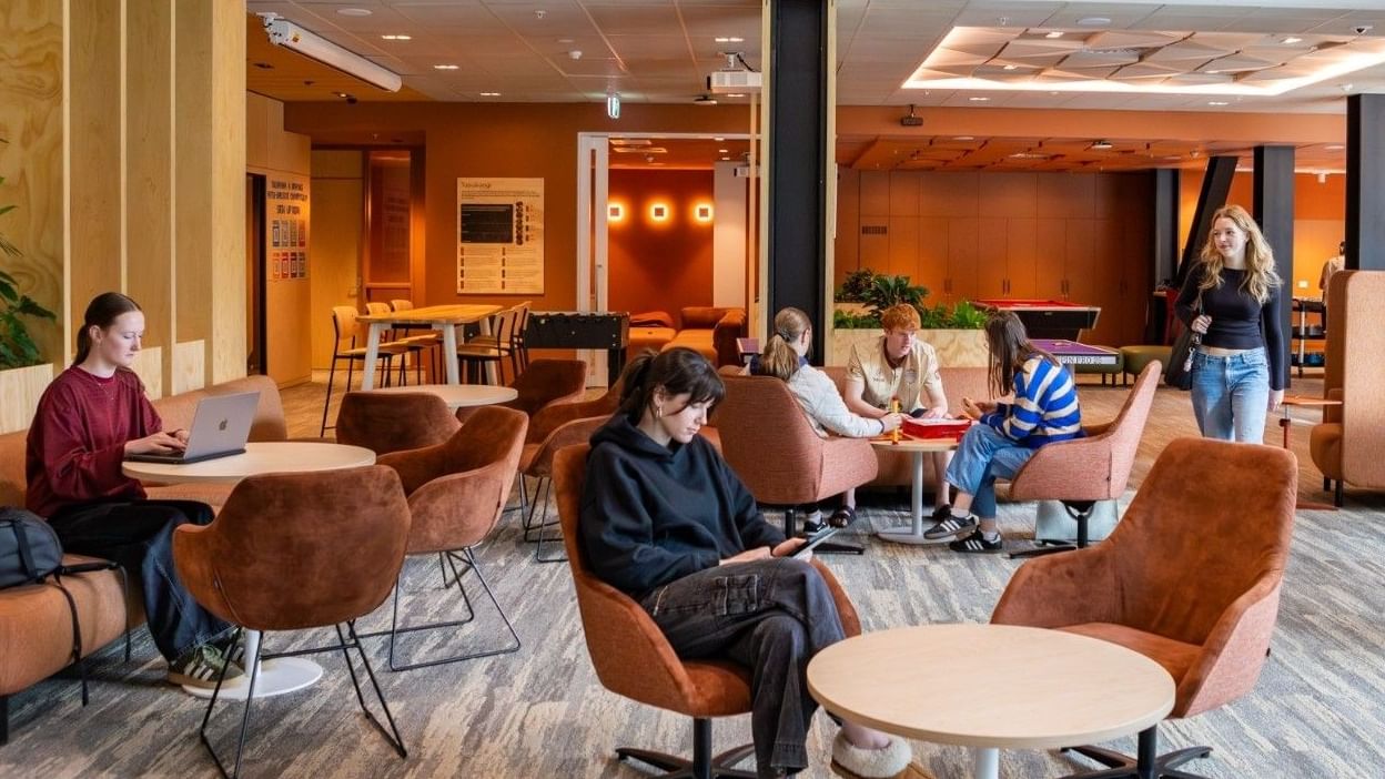 Modern communal lounge with warm wood accents, brown velvet chairs, and guests working at Tupuārangi