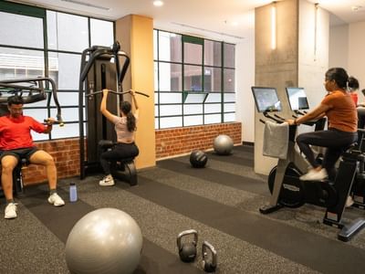 People exercising on machines in a gym with windows and brick wall.