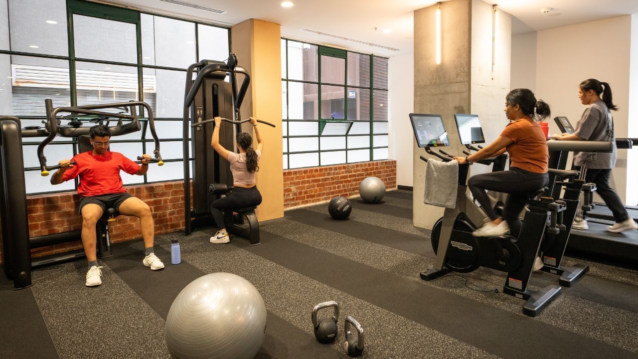 People exercising on machines in a gym with windows and brick wall.