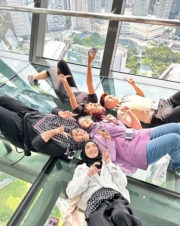 Friends lying on the glass floor, enjoying the city view from the Sky Deck at Imperial Lexis Kuala Lumpur.