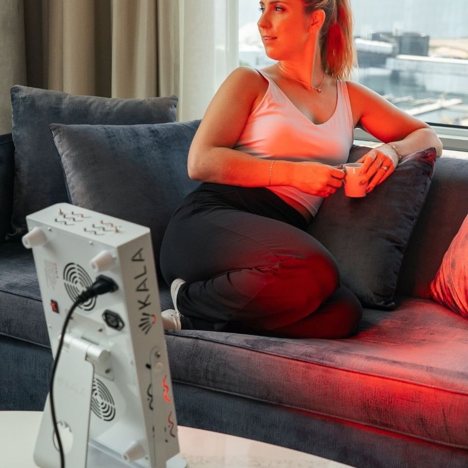 Woman relaxing with a cup on a sofa beside a glowing red light therapy device near a large window at Hotel X Toronto