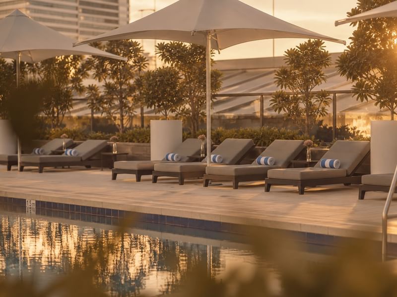 Poolside lounge chairs with white umbrellas and striped pillows at sunset with modern buildings and greenery in background.