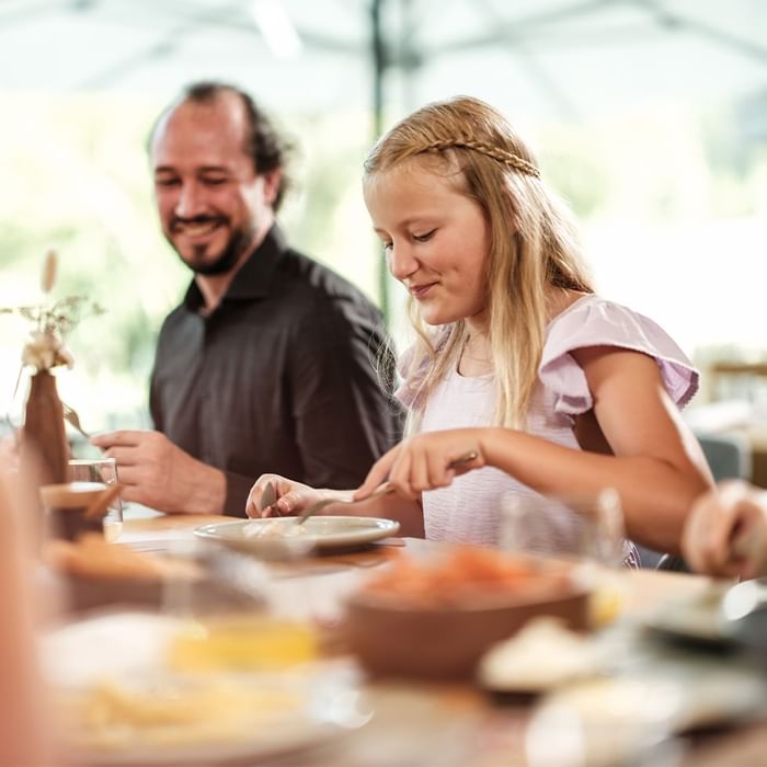 Mädchen mit geflochtenem Zopf und Mann sitzen am Tisch und essen gemeinsam.