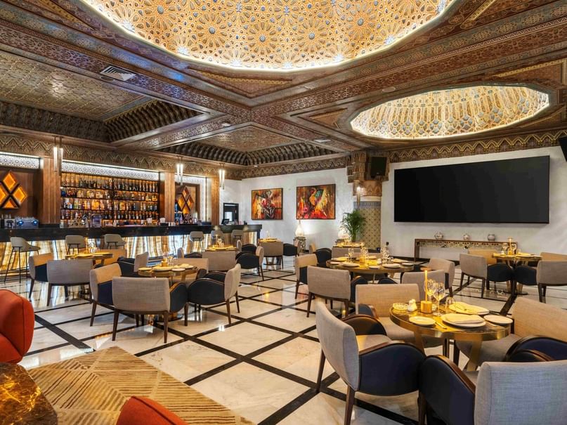 Elegant dining area with ornate ceiling, round tables, and a large wall-mounted screen at the back.