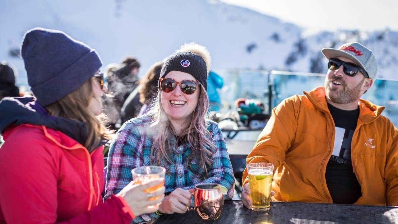 Three smiling people in winter clothes enjoying drinks at a snowy mountain setting.