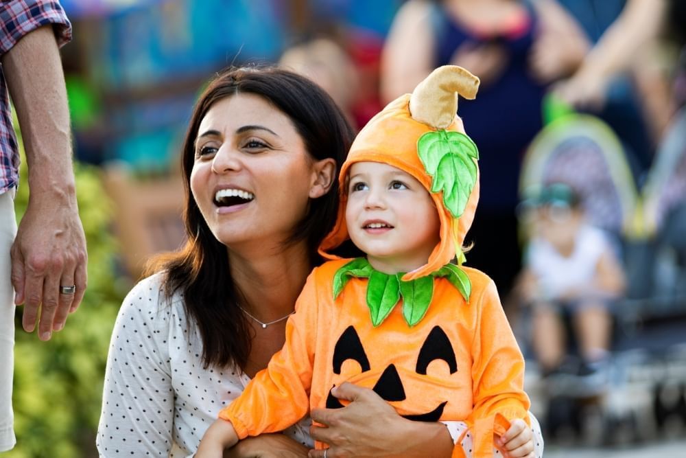 A smiling mom holds a child in a pumpkin costume outdoors.