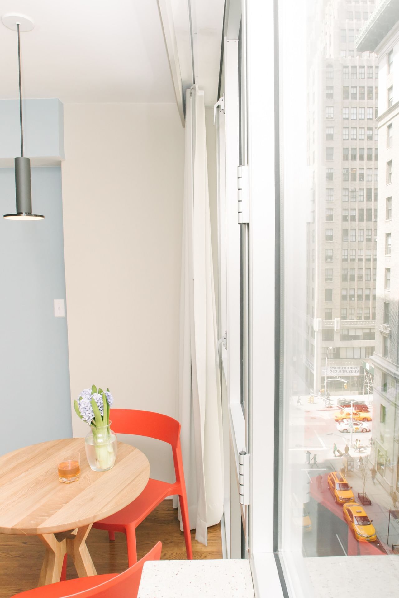 A small dining area with a round wooden table, two orange chairs, and a vase of flowers on the table, with a city view outside the window.
