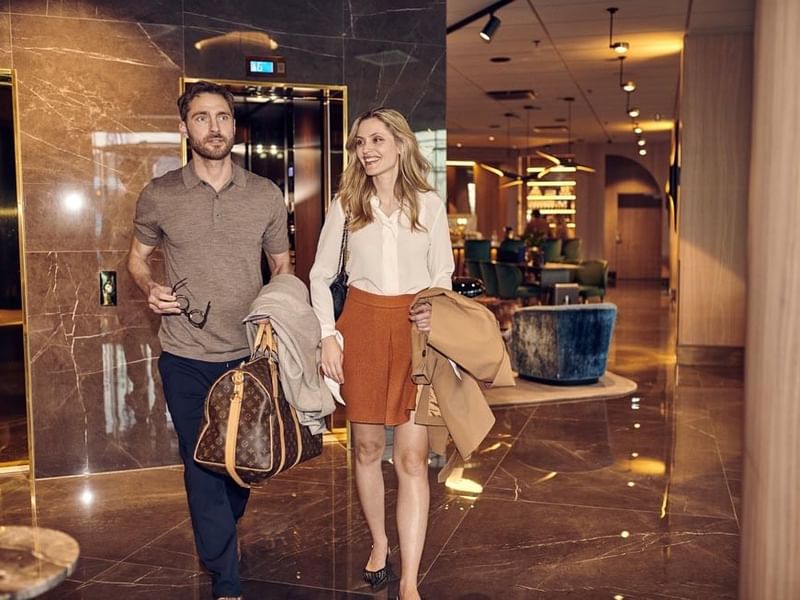 A stylish couple arrives at a luxury hotel, walking through an elegant marble lobby. The man carries designer luggage while the woman smiles, holding her coat. The hotel’s modern interior, with plush seating and warm lighting, enhances the welcoming atmos