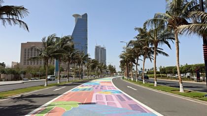 ممشى على الواجهة البحرية تصطف على جانبيه أشجار النخيل، مع أرصفة مطلية بألوان زاهية ومباني حديثة على طول كورنيش جدة والواجهة البحرية