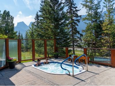 Two people relax in an outdoor hot tub at Falcon Crest Lodge, surrounded by a wooden fence, evergreen trees