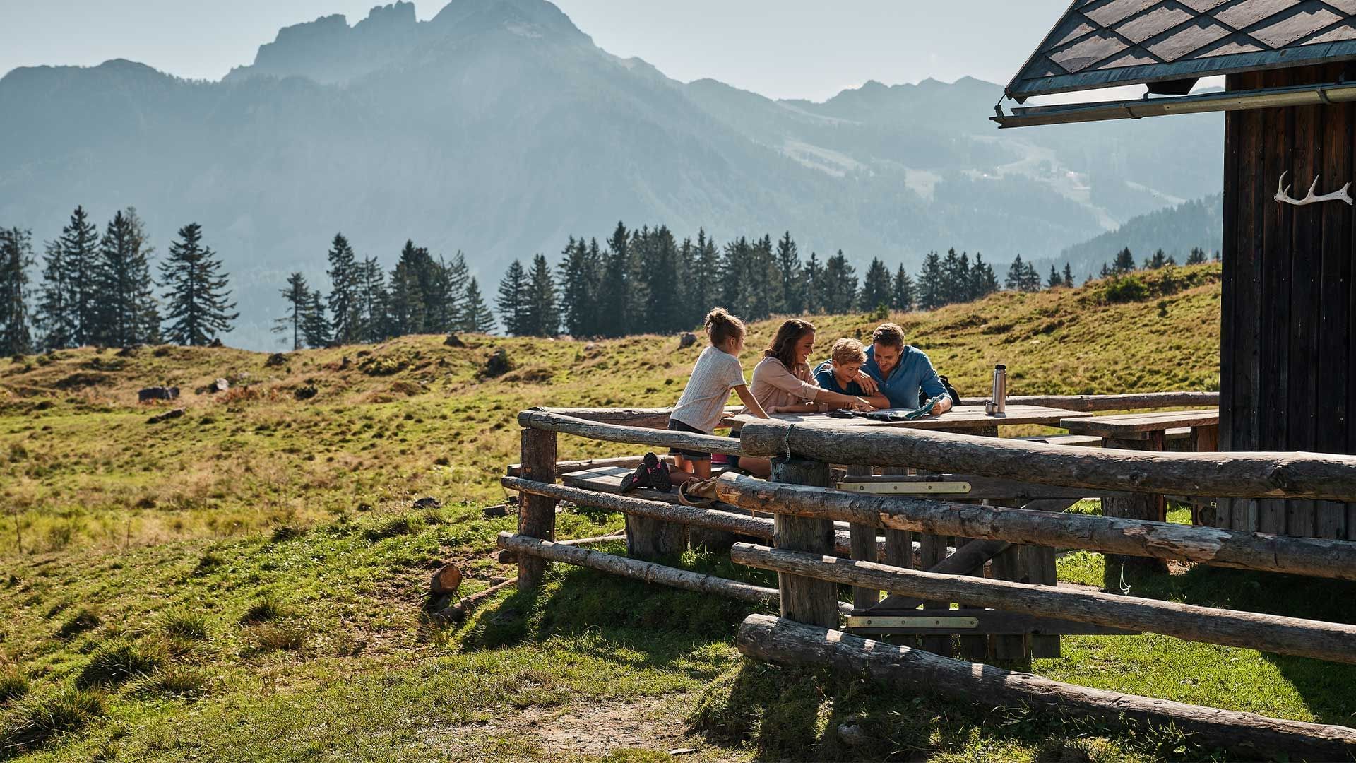 Famiglia seduta a un tavolo in legno su un prato alpino con vista sulle montagne.