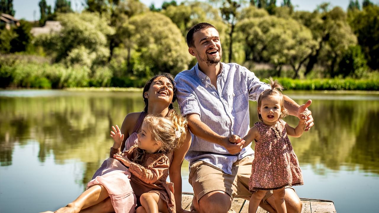 Family of four smiling and posing on a wooden dock by a lake with lush greenery.