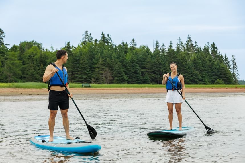 Couple paddleboarding on calm water near a lush green pine forest backdrop near Rodd Brudenell River Resort