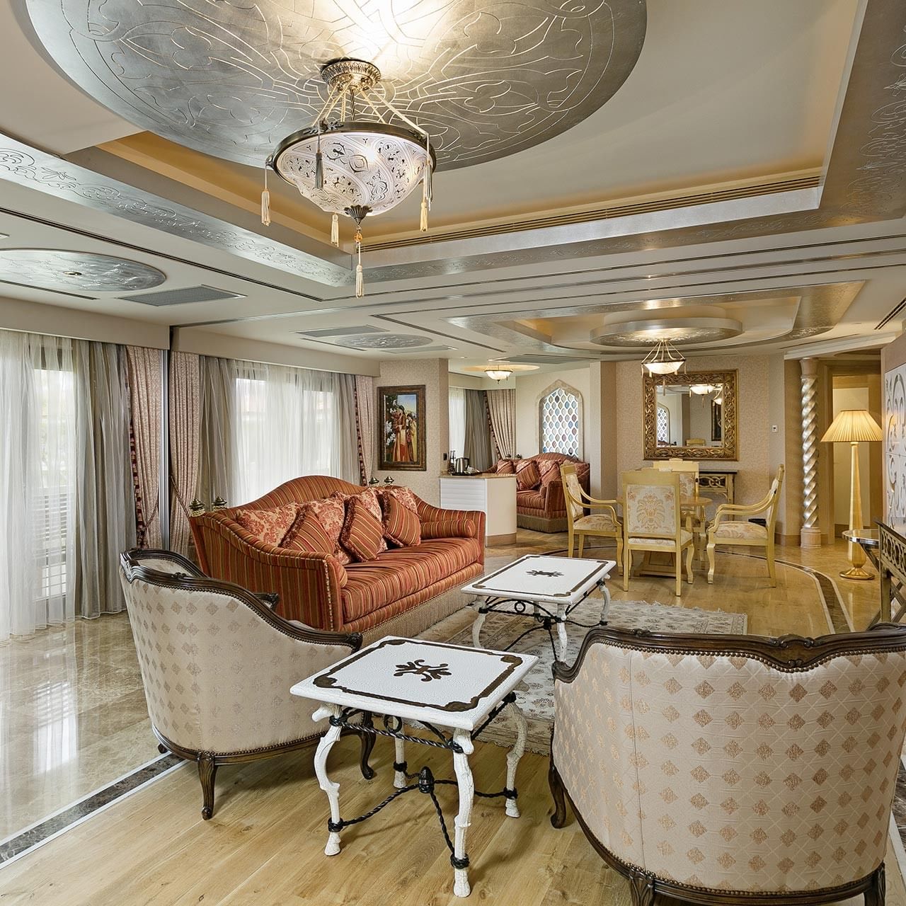 Elegantes Wohnzimmer im Lake House des Titanic Mardan Palace – mit kunstvollen Möbeln, großen Fenstern, Kronleuchter und TV auf einem Stand.