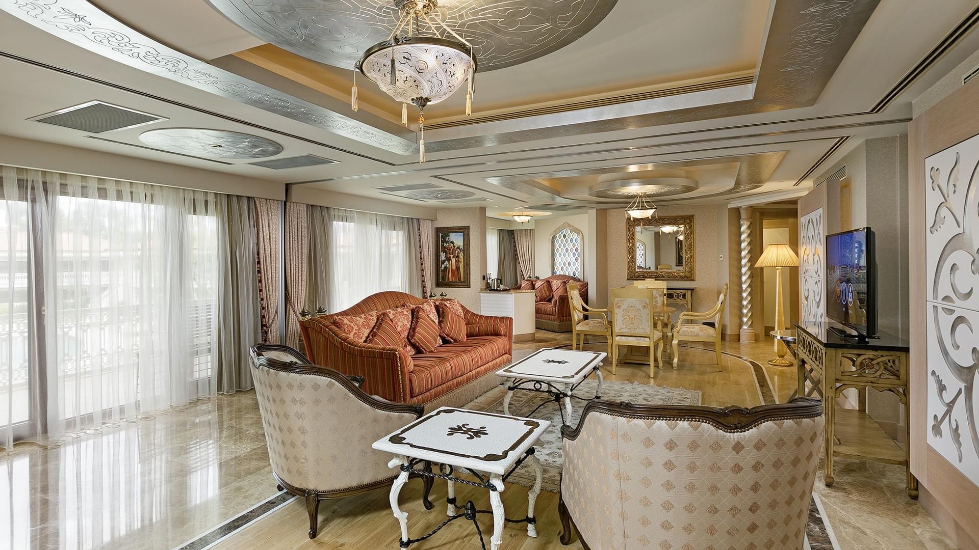 Elegantes Wohnzimmer im Lake House des Titanic Mardan Palace – mit kunstvollen Möbeln, großen Fenstern, Kronleuchter und TV auf einem Stand.