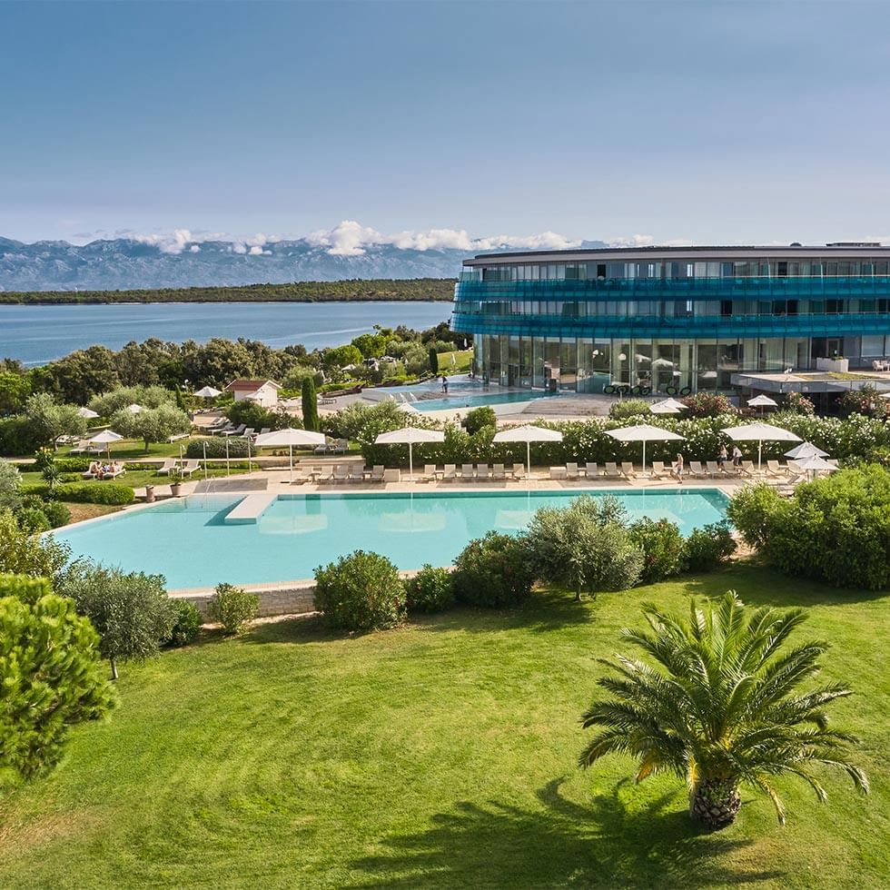 Luftaufnahme eines Resorts mit Pool, Loungebereich und Gebäuden vor einer Bergkulisse.