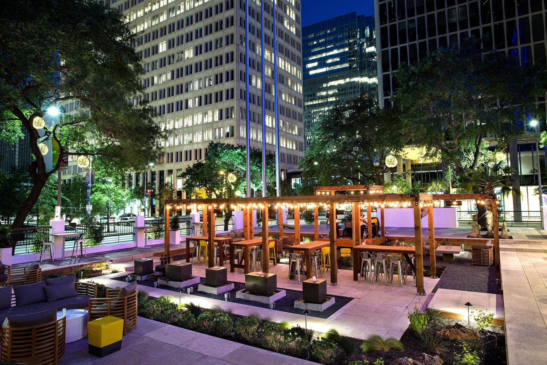 A nighttime view of an outdoor seating area with a pergola, modern furniture, and city buildings in the background.