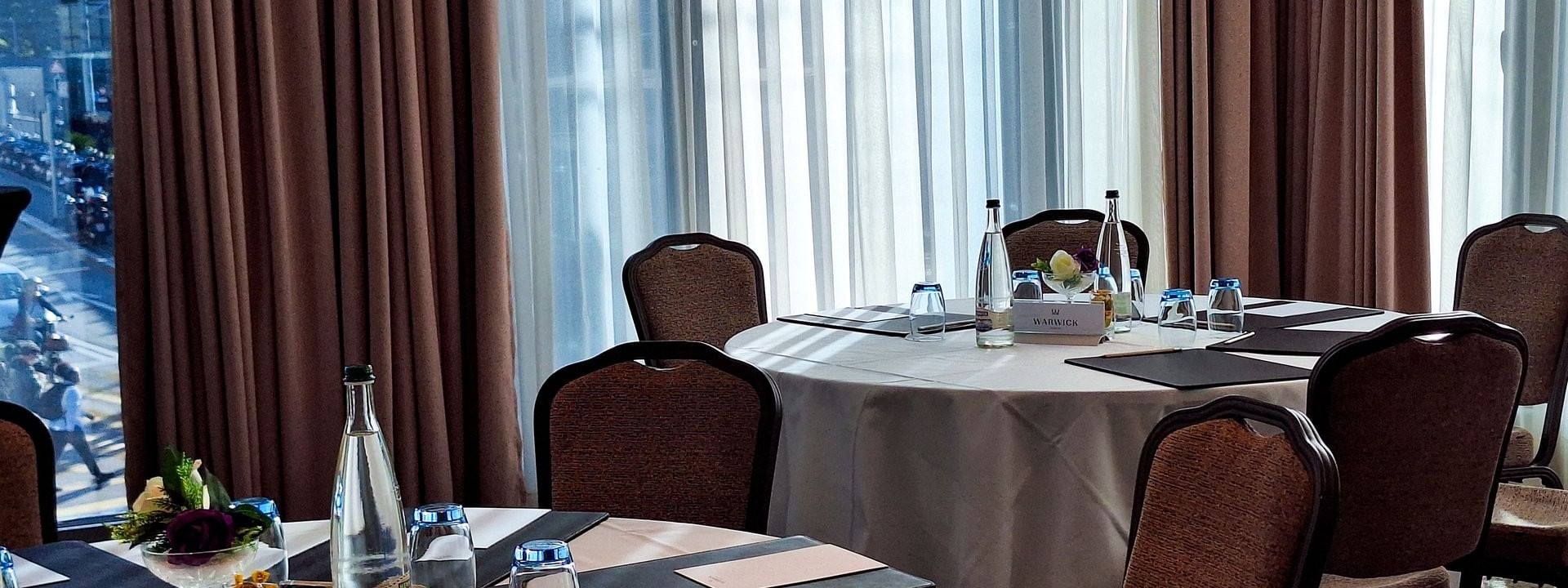Circular tables by upholstered chairs under soft daylight near a large window in Rigi + Cervin at Warwick Geneva