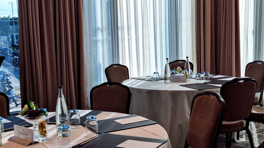 Circular tables by upholstered chairs under soft daylight near a large window in Rigi + Cervin at Warwick Geneva