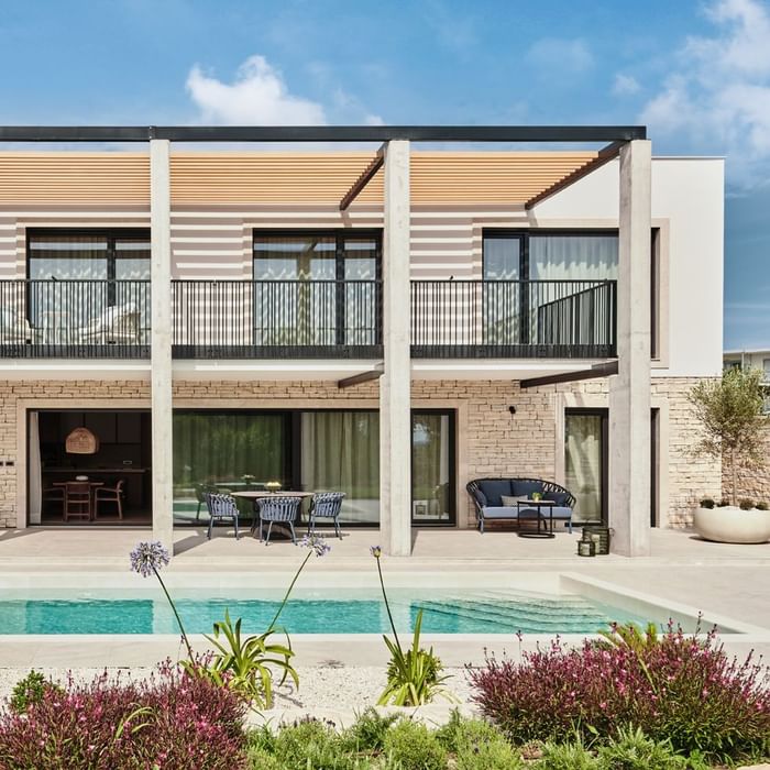 Modern two-story house with a pool, lush plants, and a patio under a blue sky at Falkensteiner Luxury Villas Punta Skala