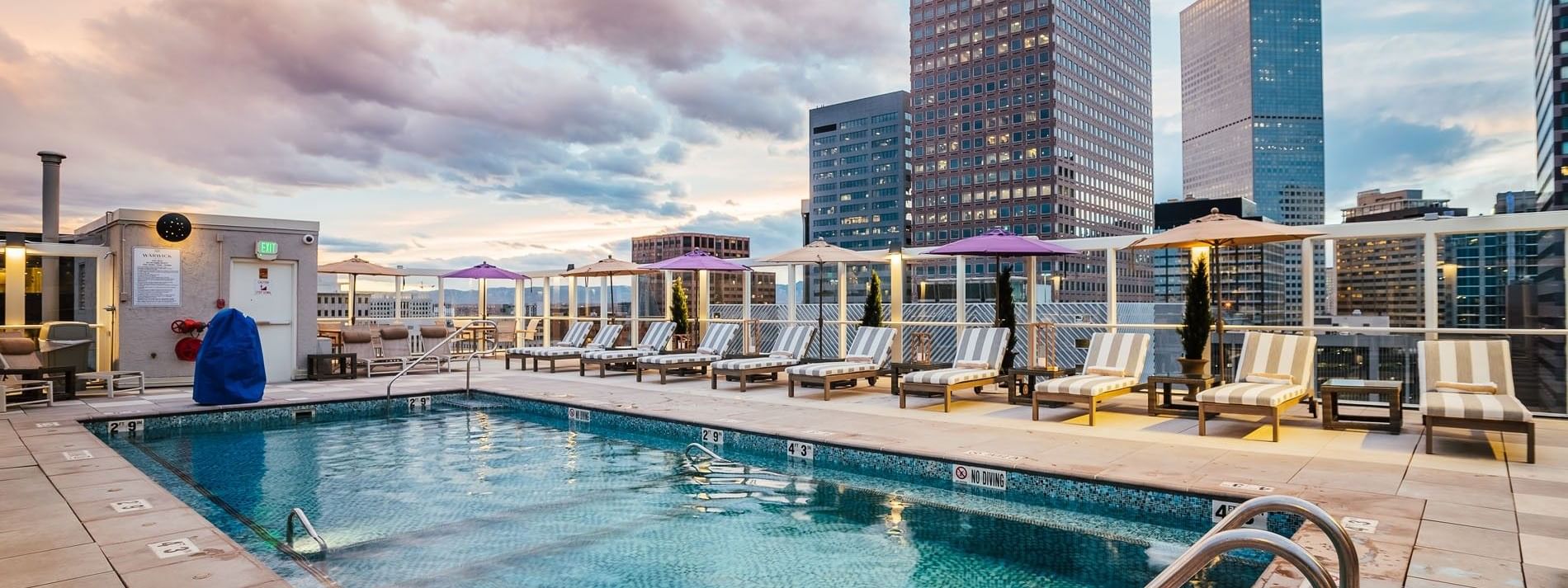 Striped lounge chairs by a blue pool under a cloudy sky near city skyscrapers in Poolside at Warwick Denver
