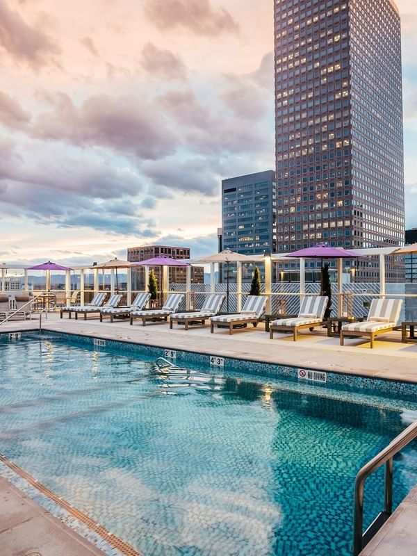 A rooftop pool with lounge chairs and umbrellas overlooking the city skyline under a cloudy sky.